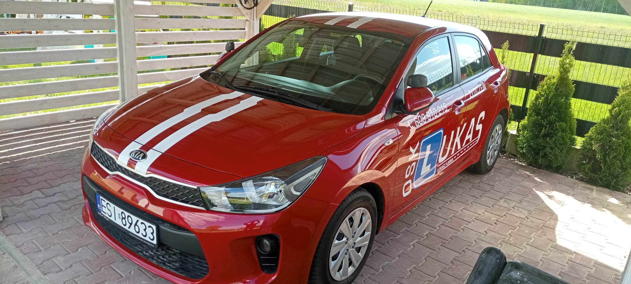 Czerwony samochód z białymi pasami i logo na boku, zaparkowany na brukowanym podjeździe, widok z przodu i boku.