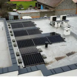 Instalacja paneli fotowoltaicznych na płaskim dachu, widoczne również jednostki klimatyzacyjne i kominy wentylacyjne, w tle zabudowania gospodarcze.