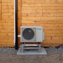 MAS Instalacje - Zewnętrzna jednostka pompy ciepła Daikin na metalowym stelażu, zamontowana przy drewnianej ścianie budynku. Widoczny fragment rury spustowej i niebieski przewód.