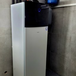 MAS Instalacje - Nowoczesna, biało-czarna pompa ciepła Daikin w pomieszczeniu z szarymi płytkami. Widoczne rury instalacyjne i niebieski zbiornik wyrównawczy. Minimalistyczny design.