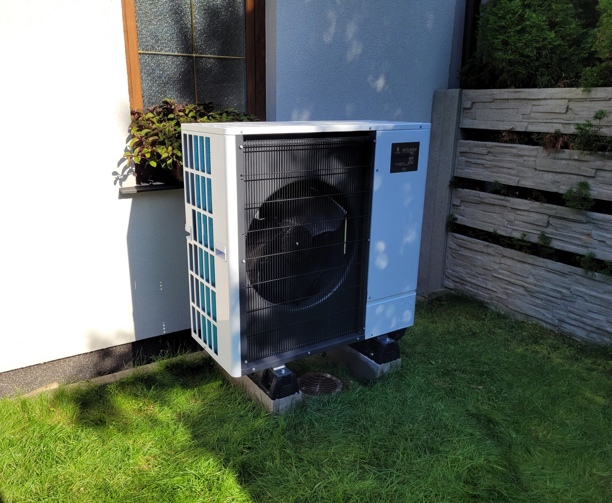 Zewnętrzna jednostka pompy ciepła Mitsubishi ustawiona na betonowych wspornikach na trawniku przy ścianie domu z oknem i ozdobnymi kwiatami w doniczce.