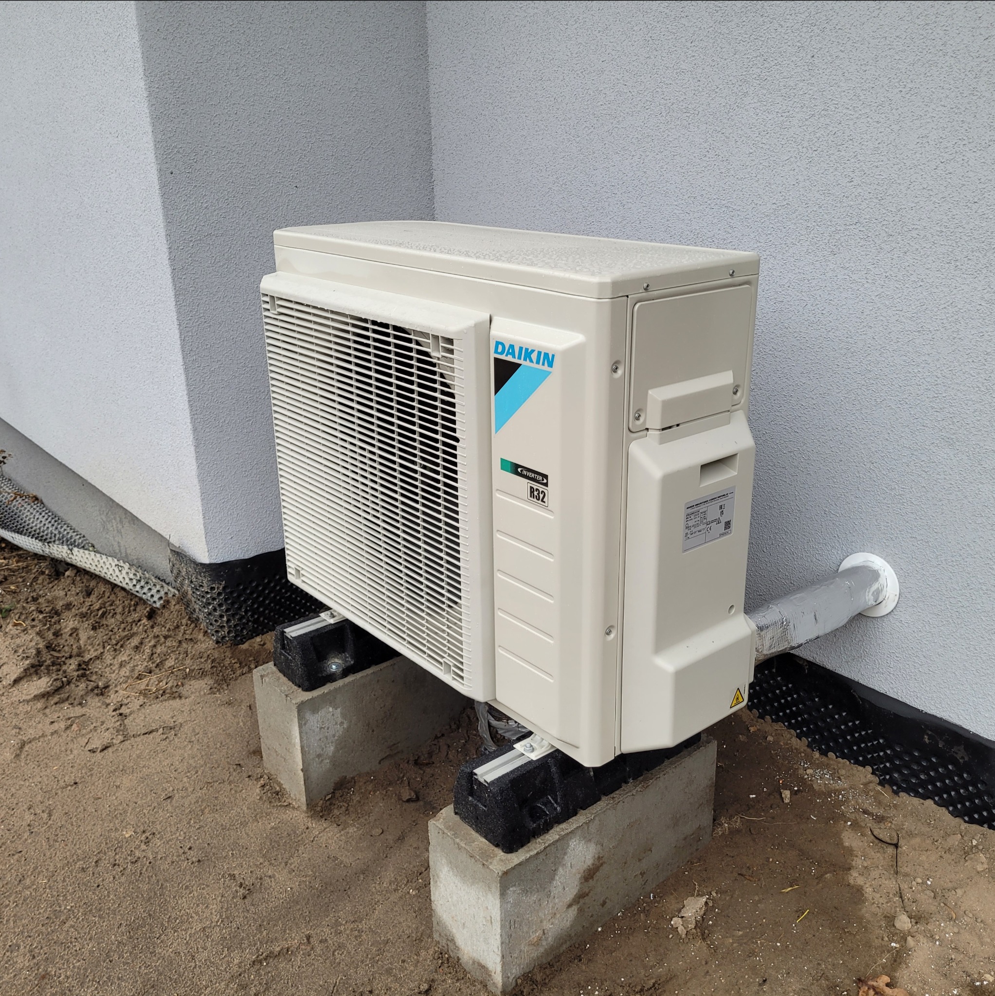 Zewnętrzna jednostka klimatyzacji Daikin zamontowana na betonowych bloczkach przy ścianie budynku z widoczną izolacją fundamentów i rurą odprowadzającą skropliny owiniętą srebrną taśmą.