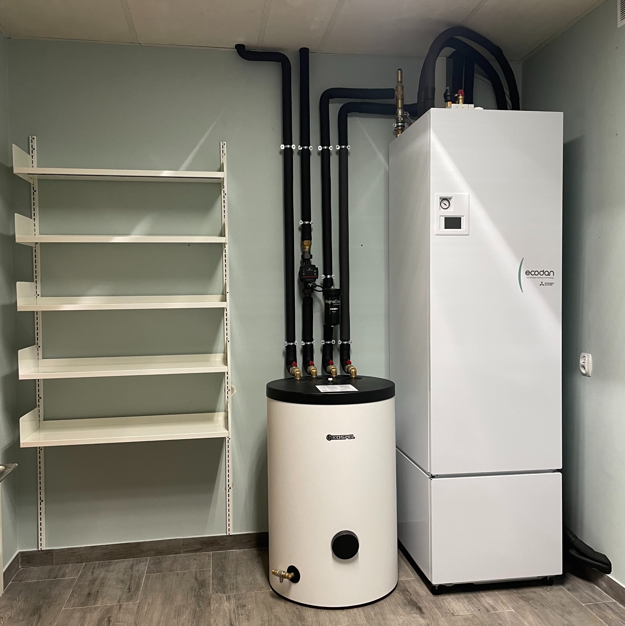 Biały zasobnik ciepłej wody Kospel i jednostka zewnętrzna Ecodan Mitsubishi Electric, połączone czarnymi rurami w pomieszczeniu z półkami na ścianie.
