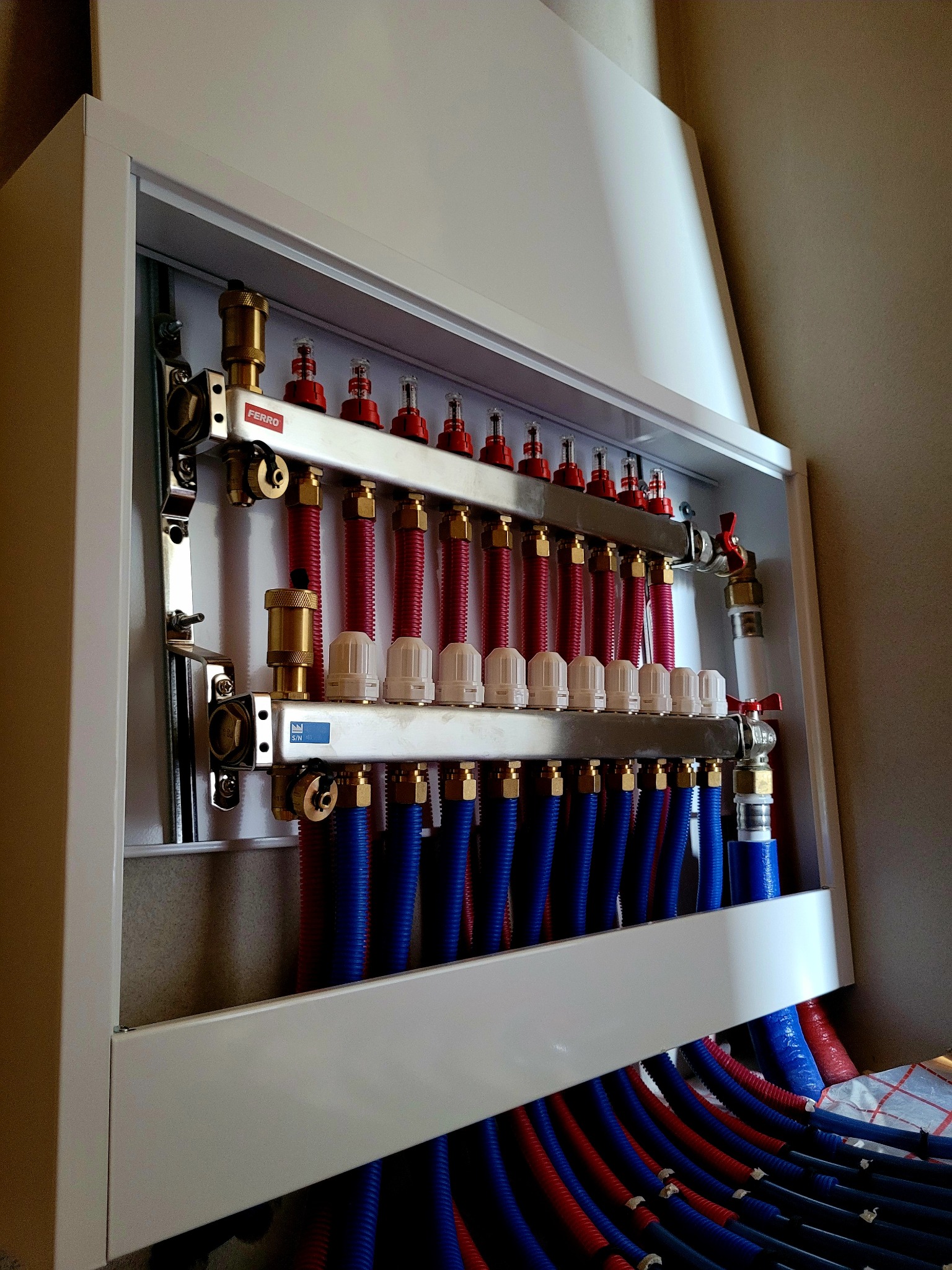 Rozdzielacz ogrzewania podłogowego Ferro w białej szafce, z widocznymi czerwonymi i niebieskimi rurami. Ujęcie z dołu, podkreślające precyzję instalacji.