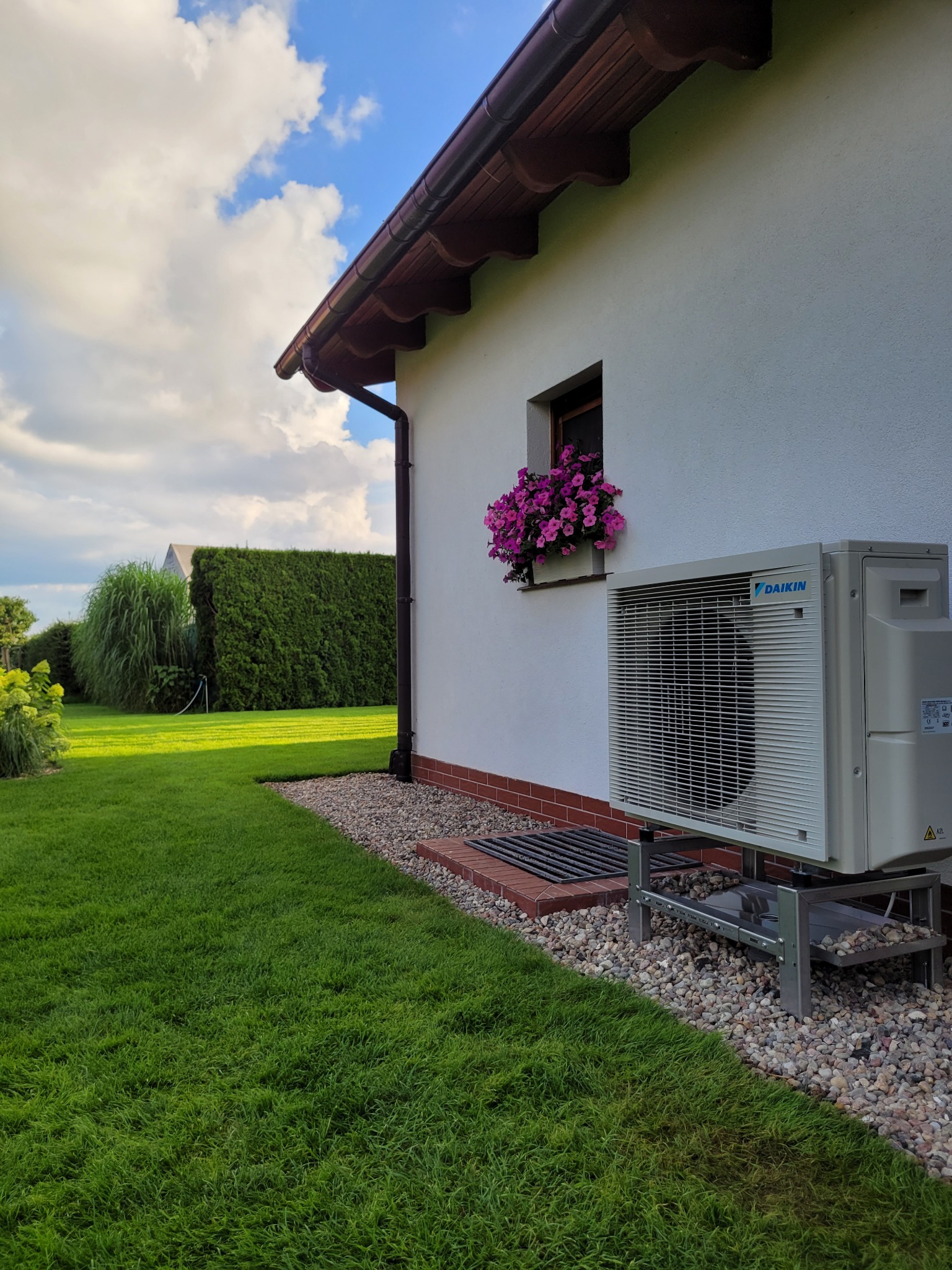 Biała pompa ciepła Daikin zamontowana na zewnątrz domu z oknem ozdobionym kwiatami. Widok na zadbany trawnik i żywopłot w słoneczny dzień.