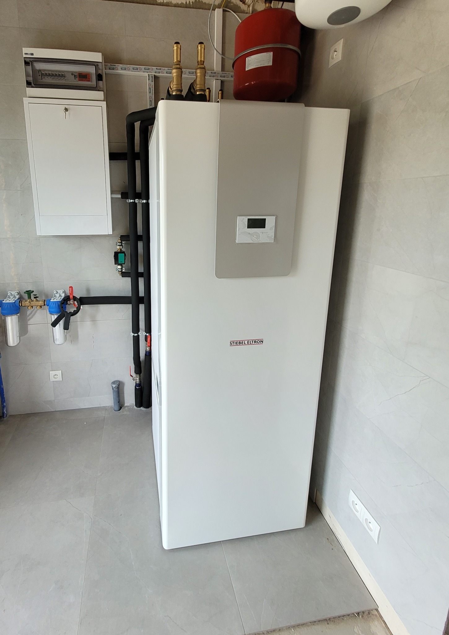 Biała pompa ciepła Stiebel Eltron z czarnymi rurami, czerwonym zbiornikiem wyrównawczym i rozdzielnicą elektryczną na tle szarych płytek w nowym pomieszczeniu.