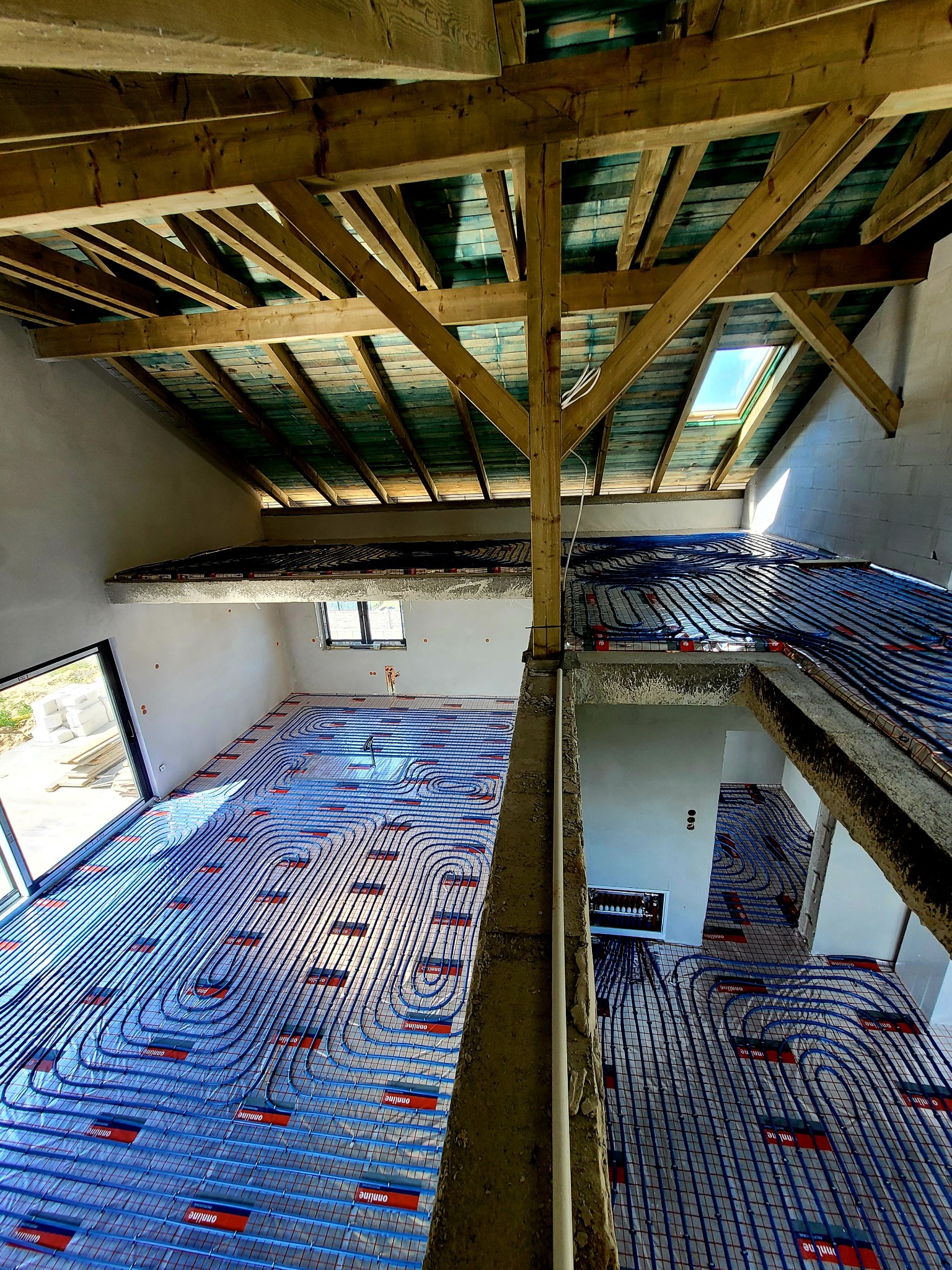 Nietypowy kadr na instalację ogrzewania podłogowego w surowym wnętrzu z drewnianą konstrukcją stropu i widocznym oknem. Niebieskie rury tworzą wzór na podłodze.