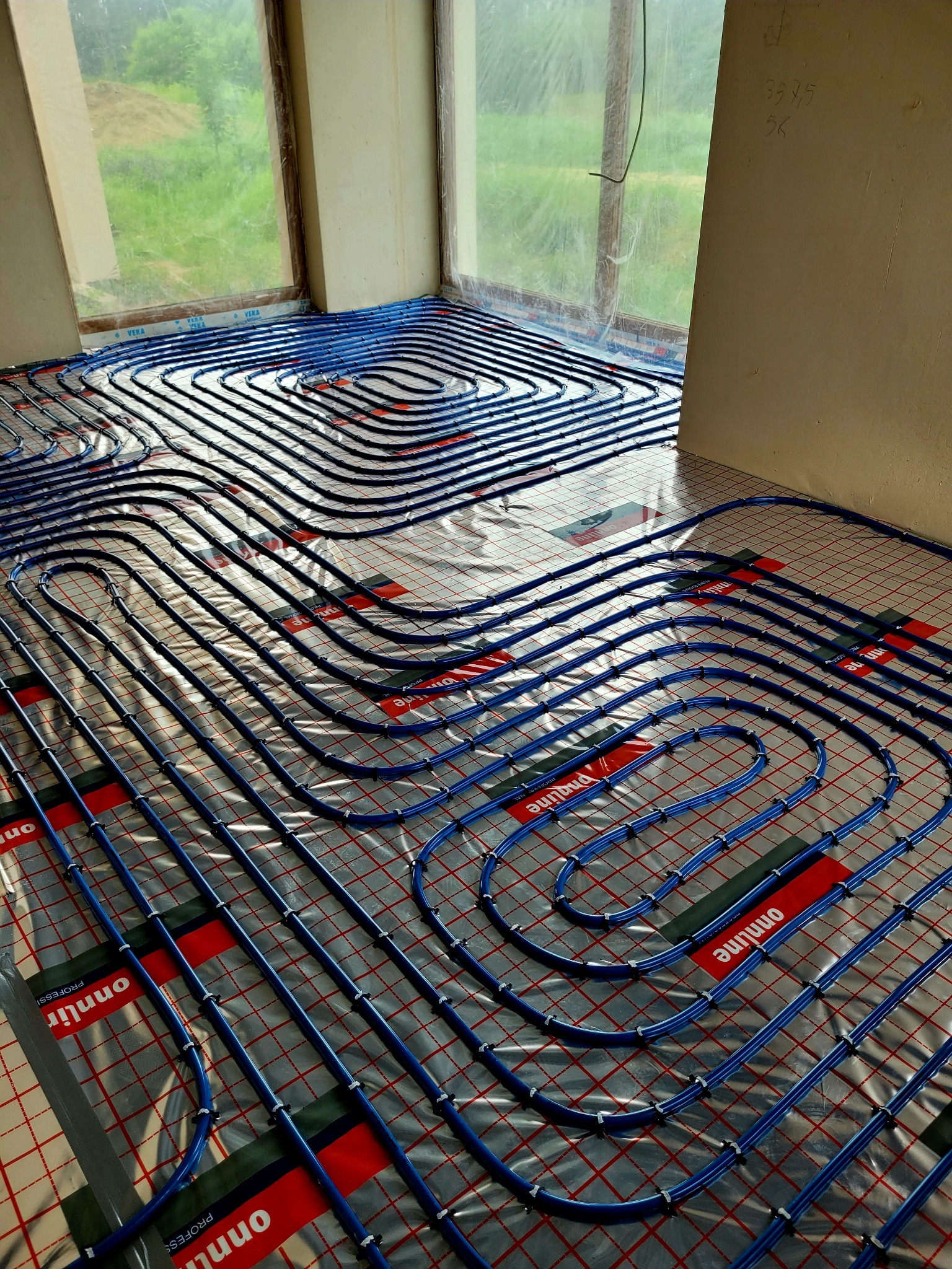 Niebieskie rury ogrzewania podłogowego ułożone spiralnie na folii z czerwoną siatką w jasnym pomieszczeniu z oknem. Widoczne mocowania rur.