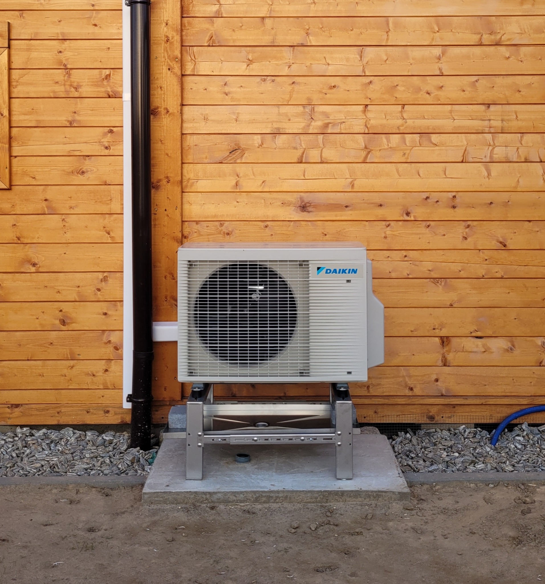Zewnętrzna jednostka pompy ciepła Daikin na metalowym stelażu, zamontowana przy drewnianej ścianie budynku. Widoczny fragment rury spustowej i niebieski przewód.