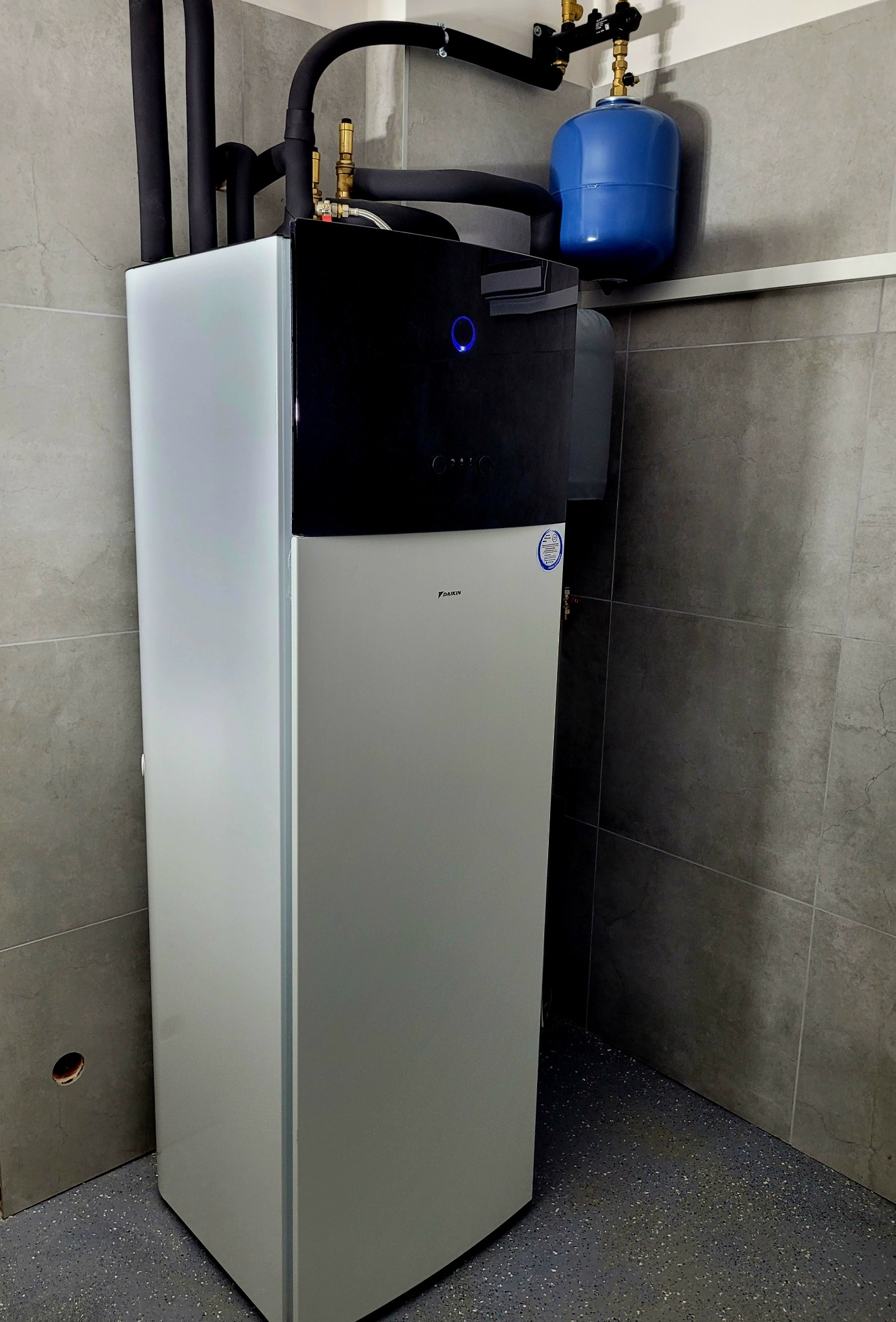 Nowoczesna, biało-czarna pompa ciepła Daikin w pomieszczeniu z szarymi płytkami. Widoczne rury instalacyjne i niebieski zbiornik wyrównawczy. Minimalistyczny design.