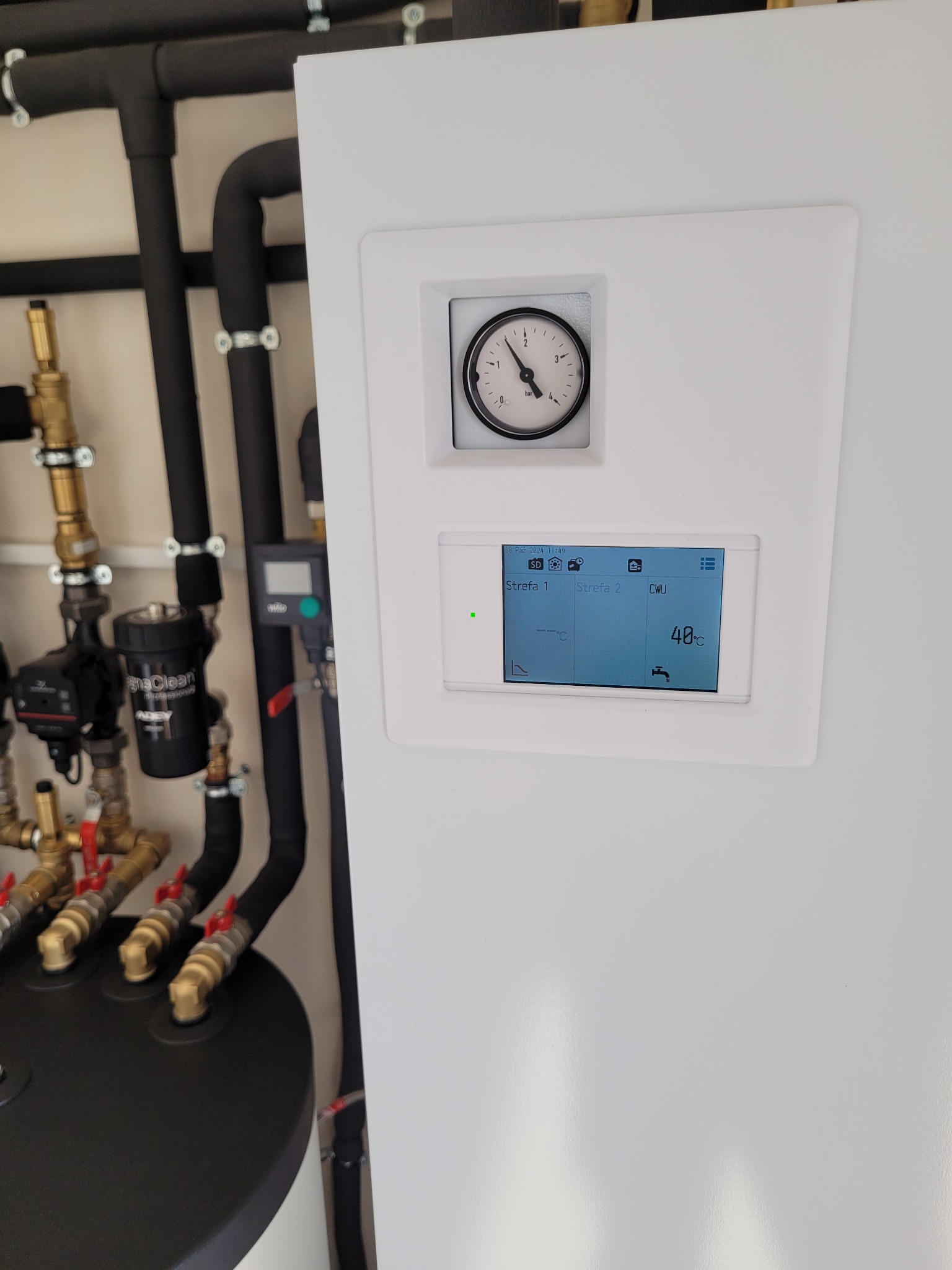 Biała obudowa pompy ciepła z widocznym manometrem i panelem sterowania wskazującym temperaturę 40°C, obok instalacja hydrauliczna z czarnymi rurami i zaworami.