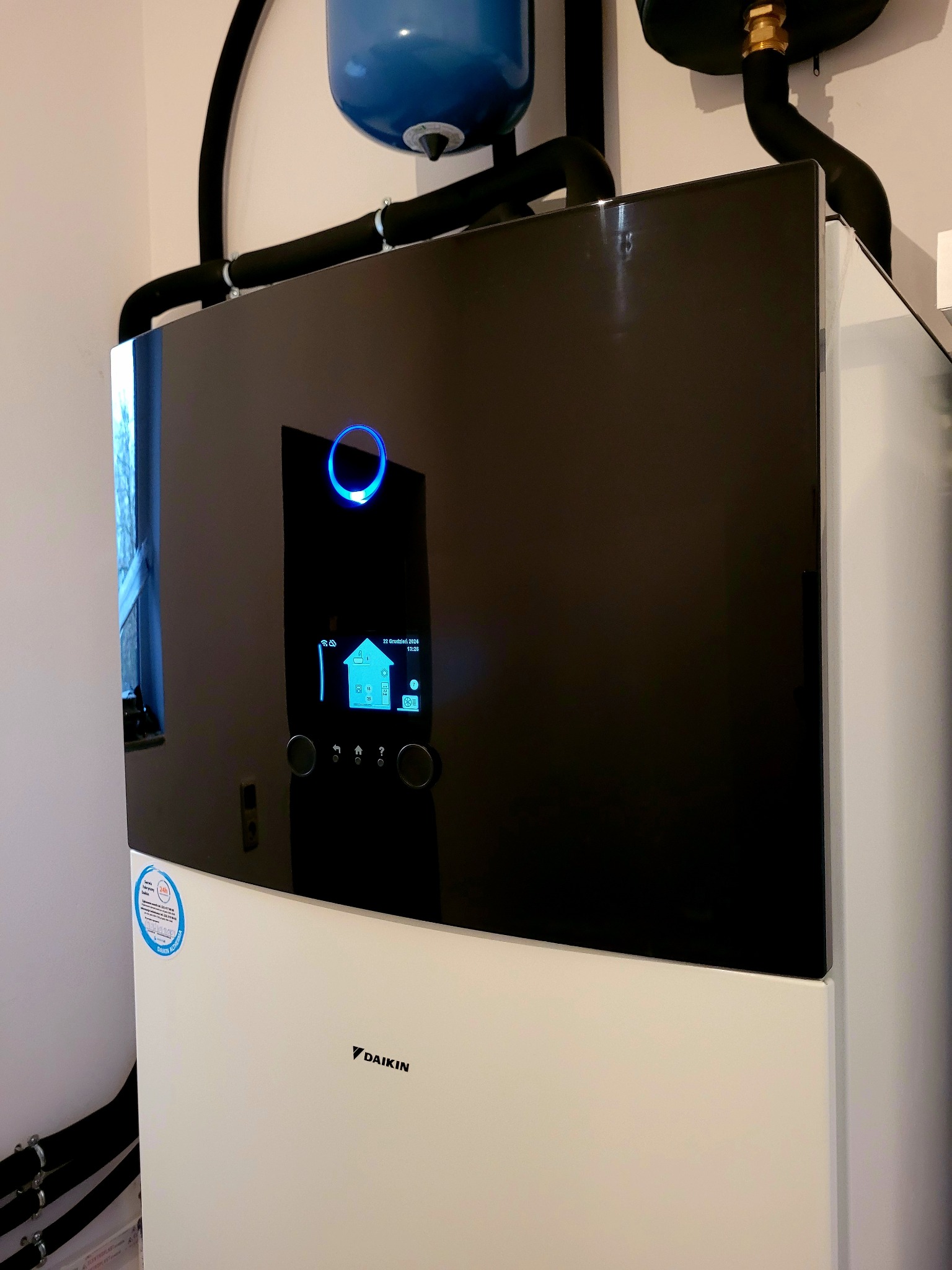 Nowoczesna pompa ciepła Daikin z czarnym panelem i niebieskim podświetleniem, widoczne rury instalacji. Kompaktowa instalacja w jasnym pomieszczeniu.