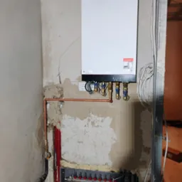 Biały piec grzewczy Viessmann zamontowany na surowej ścianie, widoczne miedziane rury, rozdzielacz ogrzewania podłogowego z czerwono-białymi pętlami, instalacja w trakcie realizacji.