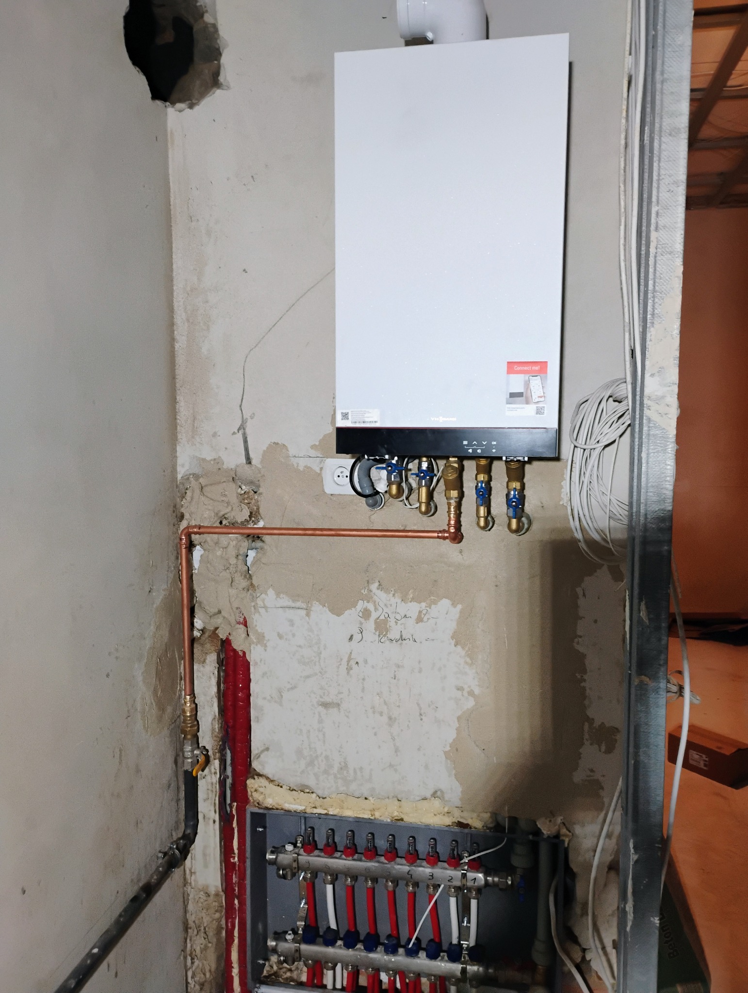 Biały piec grzewczy Viessmann zamontowany na surowej ścianie, widoczne miedziane rury, rozdzielacz ogrzewania podłogowego z czerwono-białymi pętlami, instalacja w trakcie realizacji.