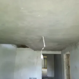 Świeżo położone, gładkie tynki na suficie i ścianach wewnątrz budynku, widoczny kabel z żarówką i fragment otwartego przejścia do innego pomieszczenia.