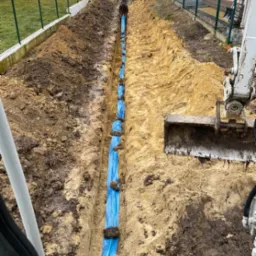 Wykop pod rury kanalizacyjne zabezpieczone niebieską folią, widok z kabiny koparki, ziemia piaszczysta, ogrodzenie z siatki po lewej stronie.