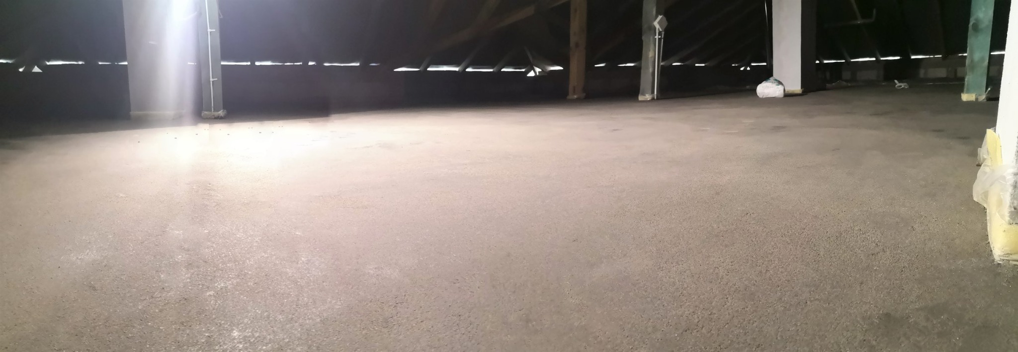 Świeżo wylana, gładka posadzka w surowym wnętrzu poddasza, z widocznymi elementami konstrukcyjnymi dachu i kolumnami wspierającymi.