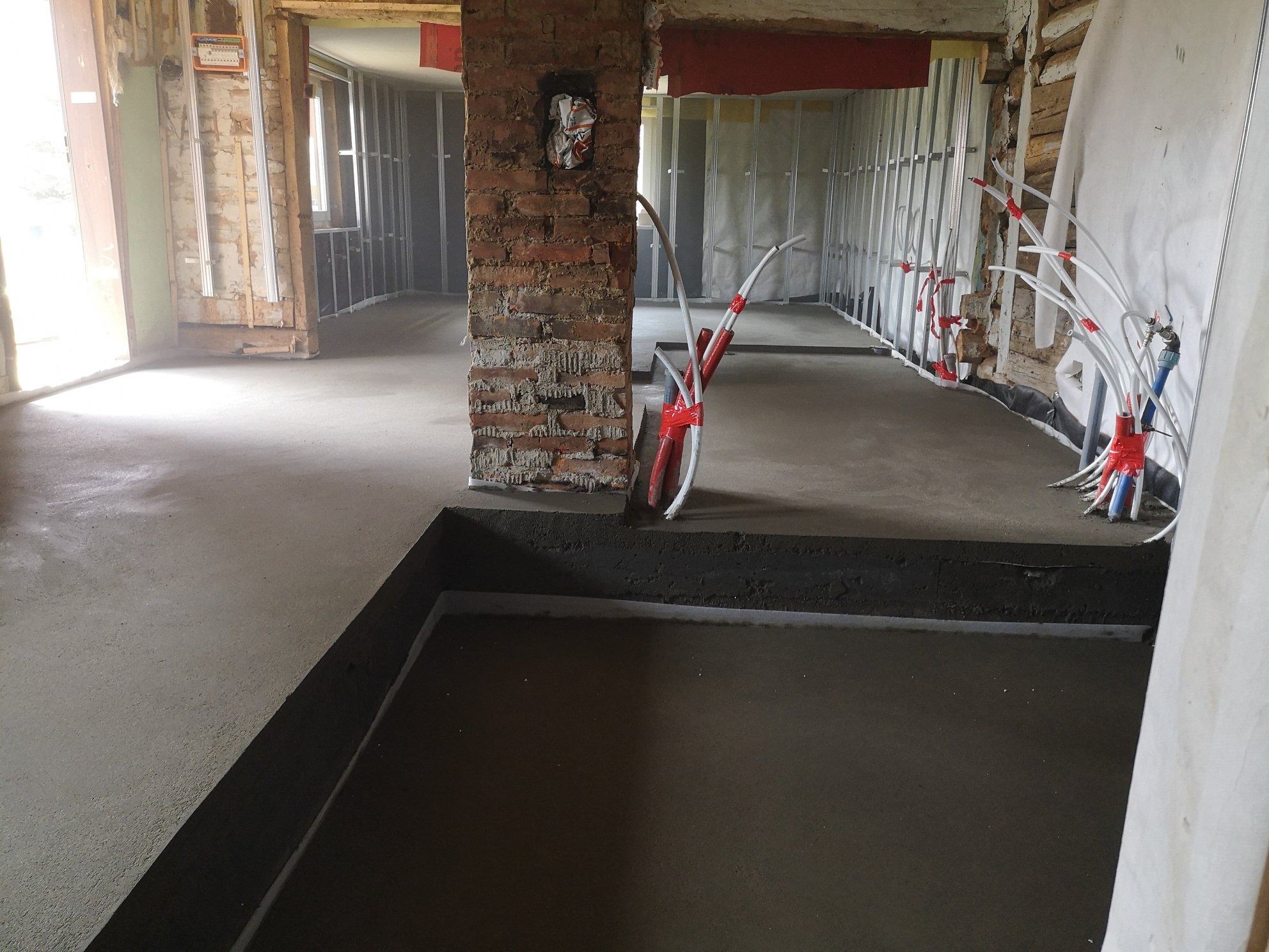 Wnętrze w trakcie remontu z wylewką podłogową, widoczne rury ogrzewania podłogowego i fragmenty ścian w stanie surowym.
