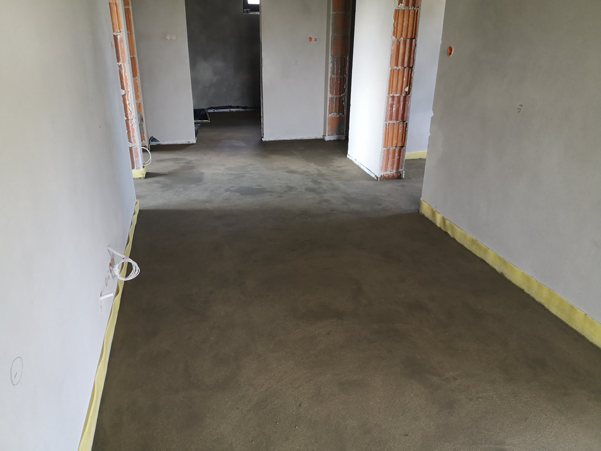 Świeżo wylana, jeszcze wilgotna posadzka w surowym stanie, z żółtą taśmą dylatacyjną przy ścianach, widoczne niedokończone ściany i fragment instalacji elektrycznej.