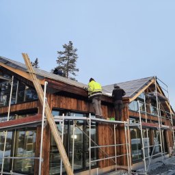 HH Happy Home - Dwóch pracowników na rusztowaniu montuje pokrycie dachu z paneli na drewnianym domu szkieletowym z dużymi oknami, widoczne narzędzia i zabezpieczenia.