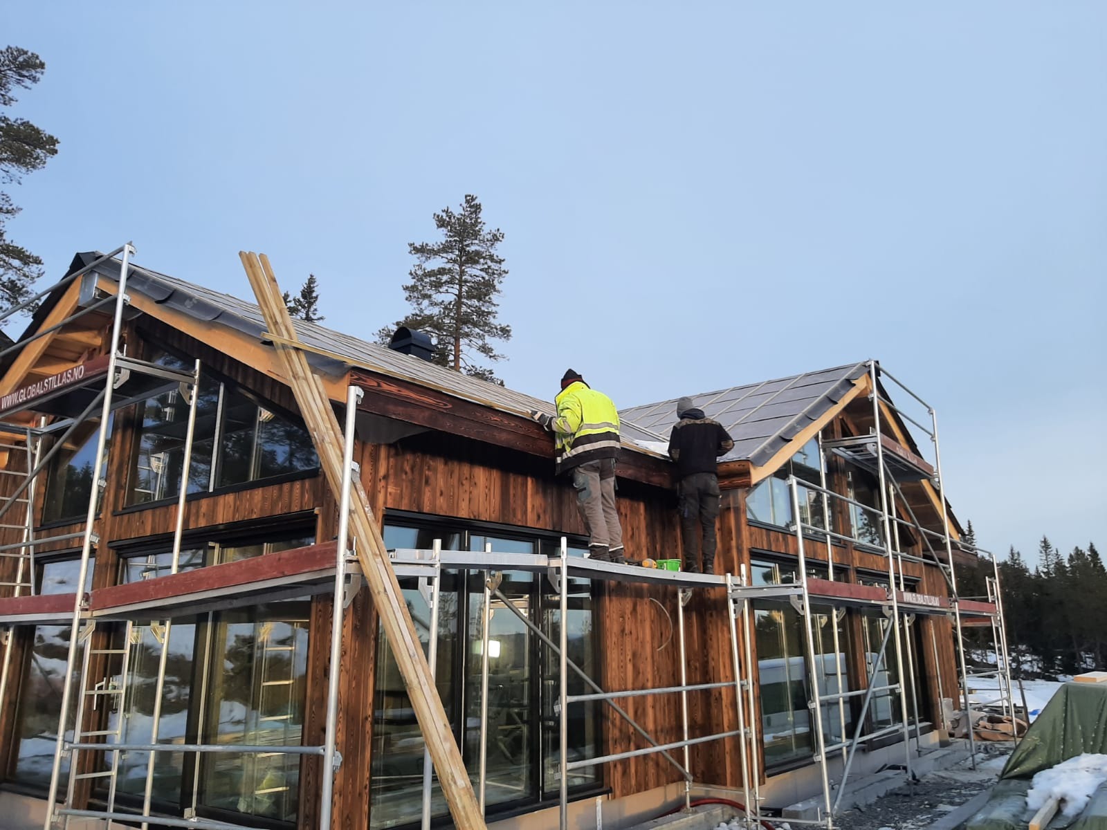 Dwóch pracowników na rusztowaniu montuje pokrycie dachu z paneli na drewnianym domu szkieletowym z dużymi oknami, widoczne narzędzia i zabezpieczenia.