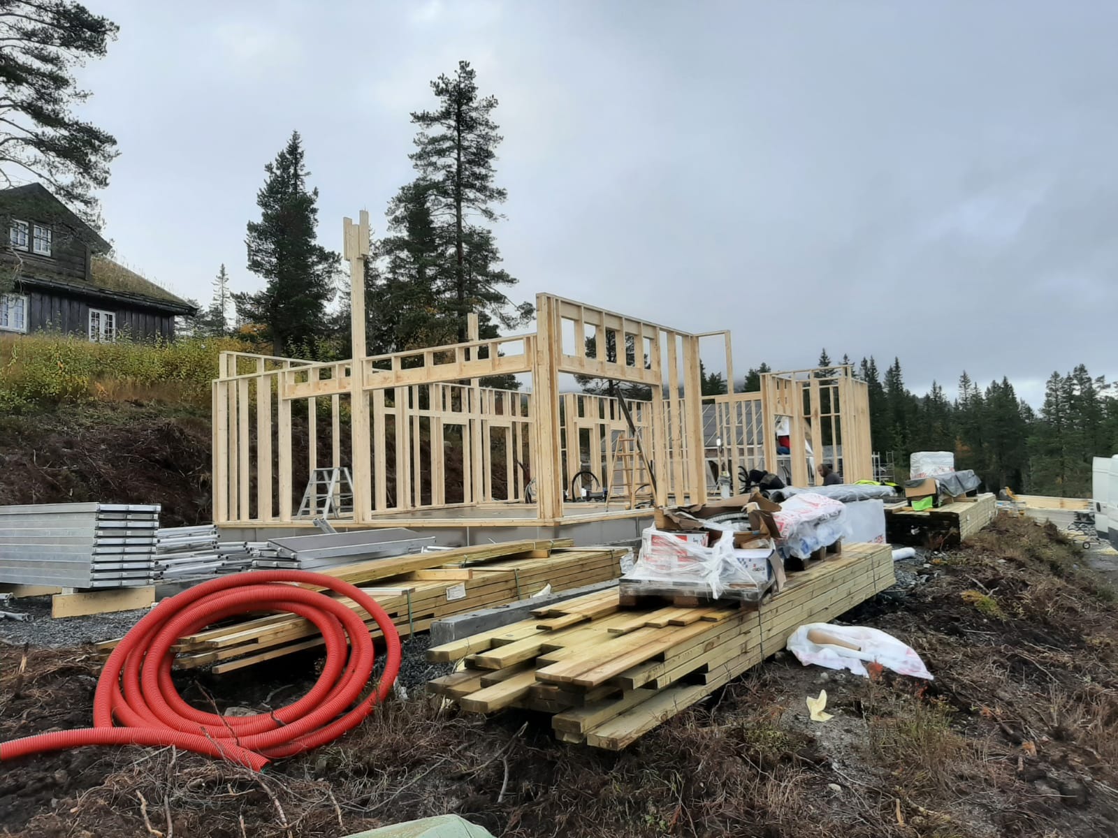 Wznoszenie drewnianej konstrukcji domu szkieletowego na fundamencie, widoczne materiały budowlane i czerwony zwój rur, w tle dom z zielonym dachem i las.