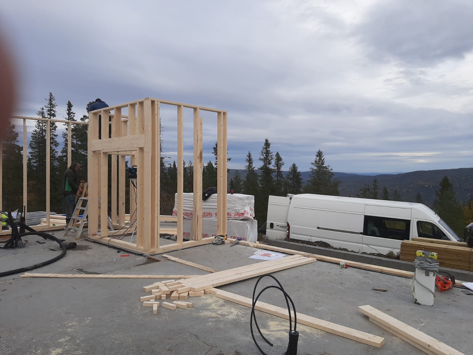 Konstrukcja szkieletowa domu w trakcie budowy, widoczne drewniane ramy ścian, pracownicy na drabinie, materiały budowlane, narzędzia i biały samochód dostawczy na tle zalesionych wzgórz.