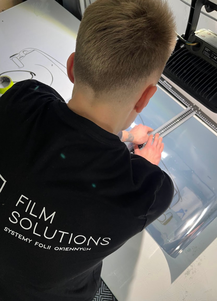 Pracownik firmy Film Solutions przycina folię okienną na stole roboczym, używając linijki i nożyka, widoczny z tyłu w czarnej koszulce z logo firmy.