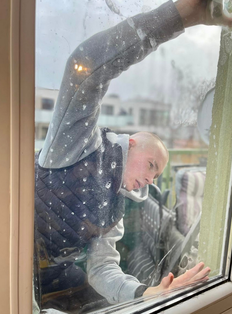 Młody mężczyzna w szarej bluzie i czarnej kamizelce czyści okno od wewnątrz, widoczne krople wody i piany na szybie, z zewnątrz widok na budynki.