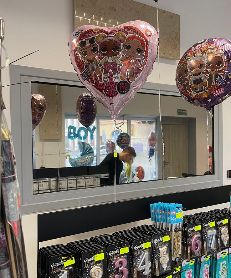 Sklep z artykułami imprezowymi: balony foliowe z postaciami LOL Surprise w kształcie serca i kuli, świeczki urodzinowe cyfry na półce, dekoracje na baby shower widoczne w odbiciu szyby.
