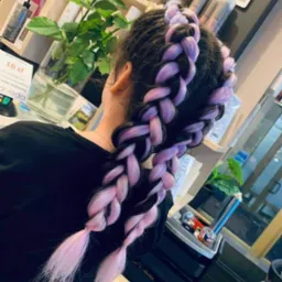 Uczesanie z grubych, fioletowo-r&oacute;żowych warkoczy na ciemnych włosach, upiętych wok&oacute;ł głowy. Widoczne produkty do pielęgnacji włos&oacute;w i dekoracyjna r&oacute;ża w wazonie w tle.