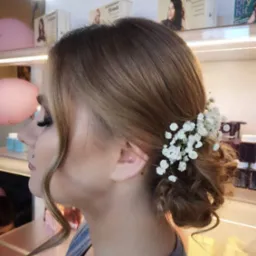 Upięcie ślubne z wpiętymi białymi drobnymi kwiatami, widok z profilu na jasnobrązowe włosy z blond pasemkami, na tle kosmetyki Kevin Murphy.