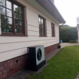 Zewnętrzna jednostka pompy ciepła typu monoblok, zainstalowana przy ścianie domu z beżową elewacją i brązowymi oknami, widoczna z boku na tle trawnika.