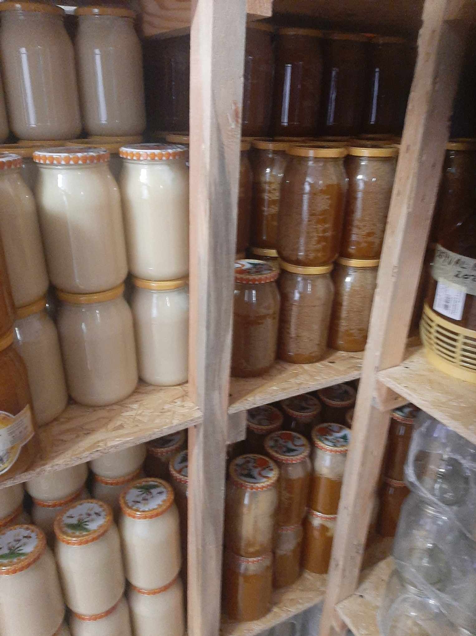 Półki regału wypełnione słoikami z miodem o różnej barwie, od jasnego kremowego do ciemnego bursztynowego, niektóre słoiki z dekoracyjnymi wieczkami, ustawione w surowym, drewnianym regale.