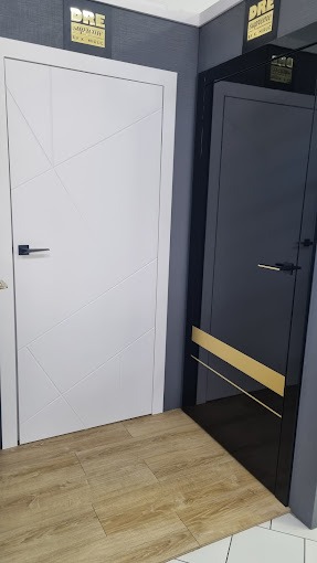 Ekspozycja w salonie: białe drzwi z geometrycznym wzorem i czarne drzwi z poziomym złotym paskiem, oba modele DRE Supreme.