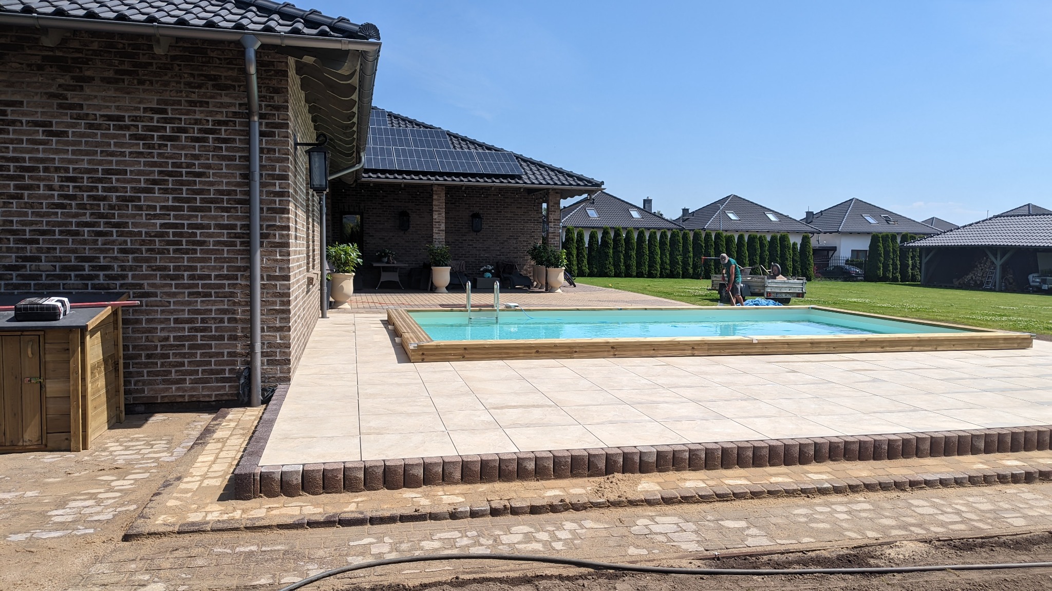 Nowoczesny basen z jasną wodą i drewnianą obudową, otoczony płytami tarasowymi w kolorze beżowym, z widocznymi fragmentami niedokończonej nawierzchni z kostki brukowej i domem z panelami...