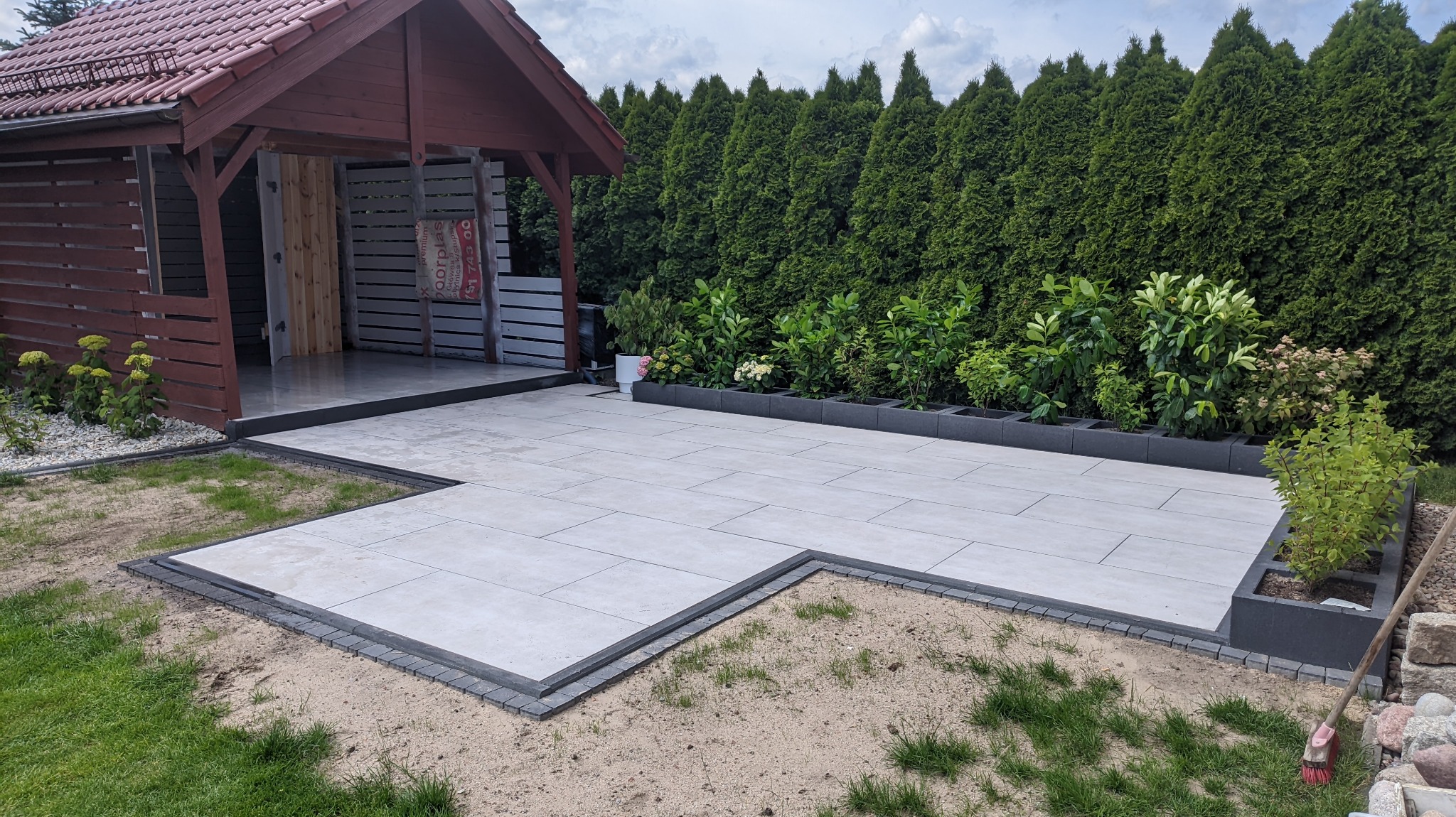 Wykończony taras z jasnych płyt w Krępie Słupskiej, obrzeża z ciemnej kostki, rabaty z roślinami w donicach, altana w tle.