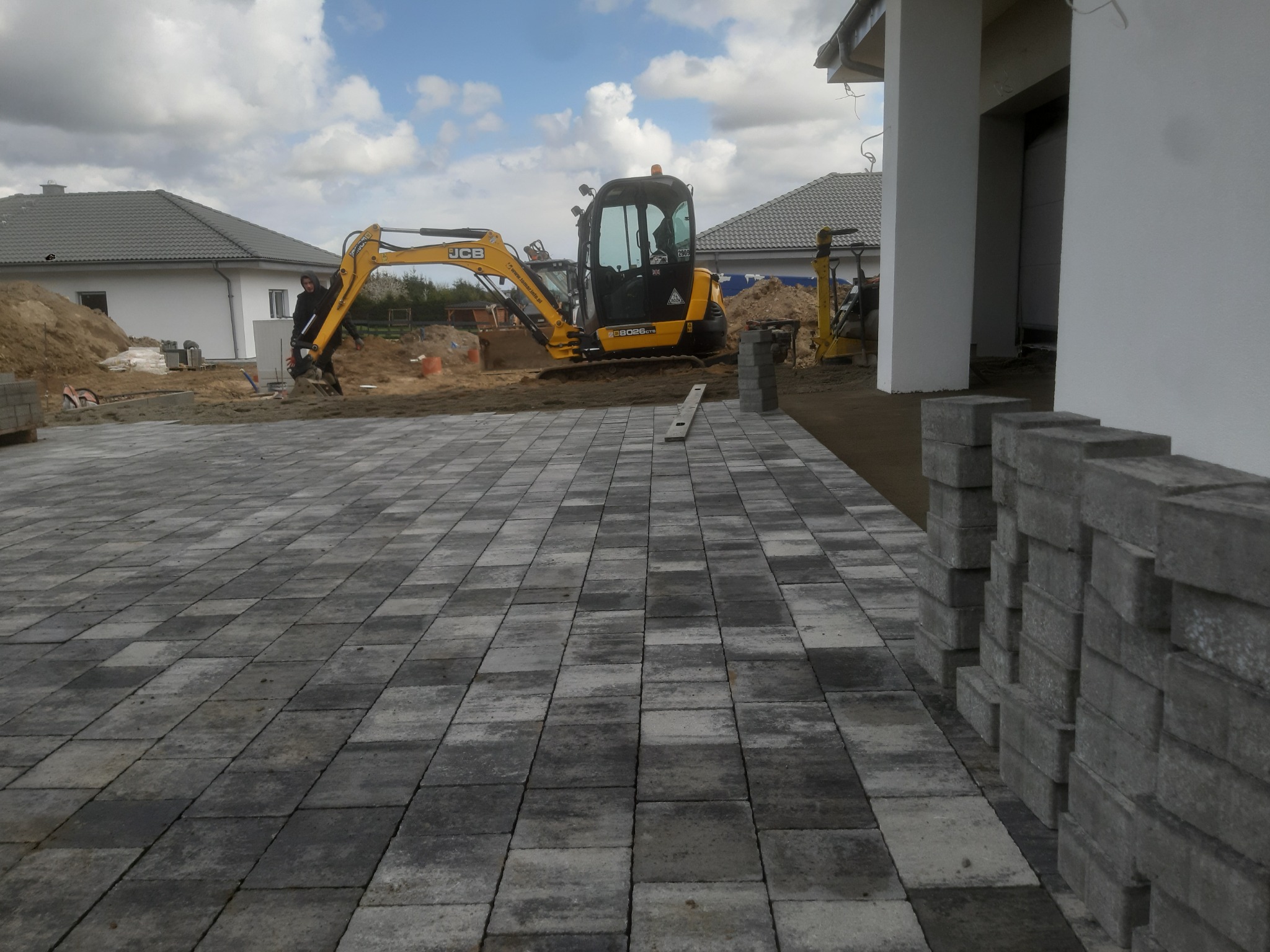 Układanie kostki brukowej w kolorze szarości, z żółtą koparką JCB w tle i stosem kostki po prawej stronie, widok na budowę domu jednorodzinnego.