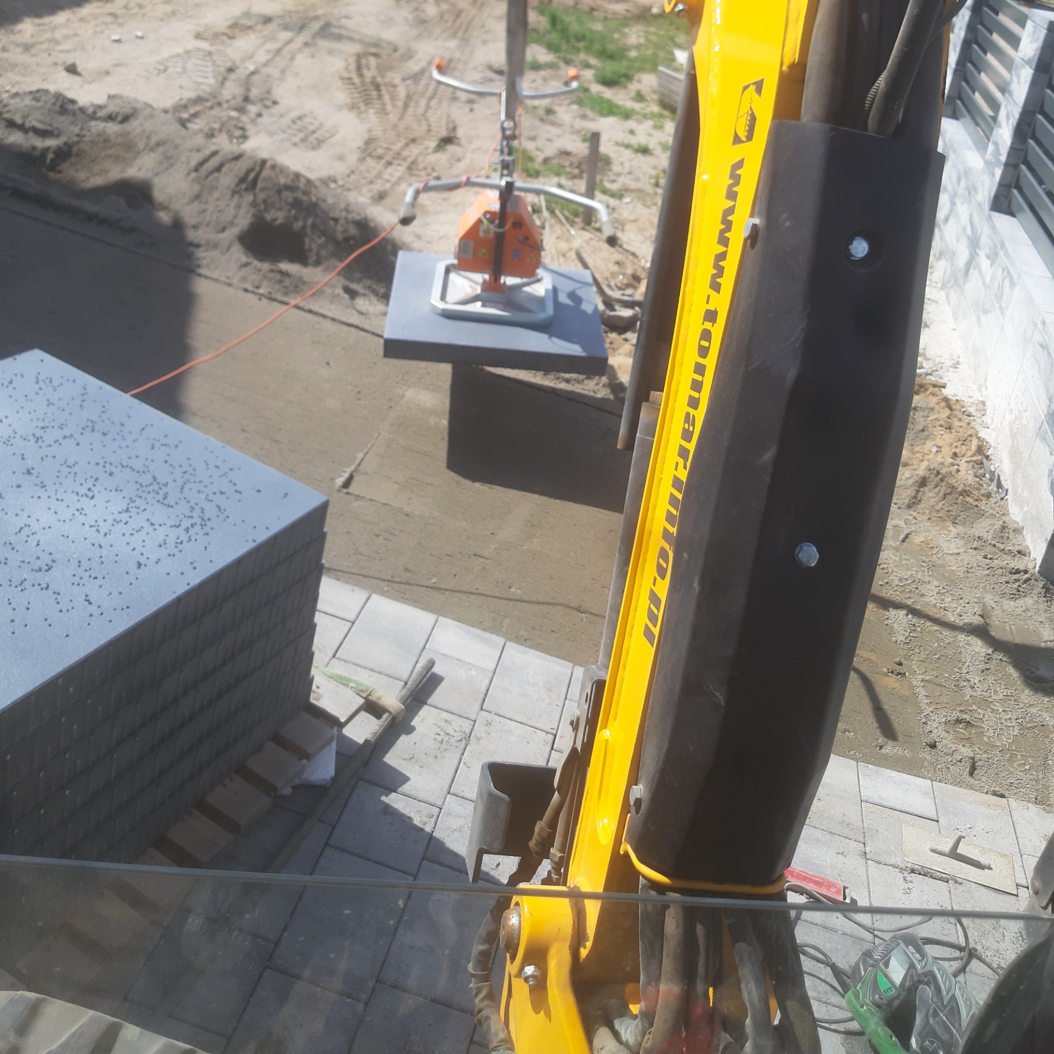 Żółty podnośnik próżniowy przenosi szarą płytę chodnikową nad świeżo ułożoną kostką brukową. W tle stos płyt chodnikowych i przygotowany teren pod kolejne płyty.