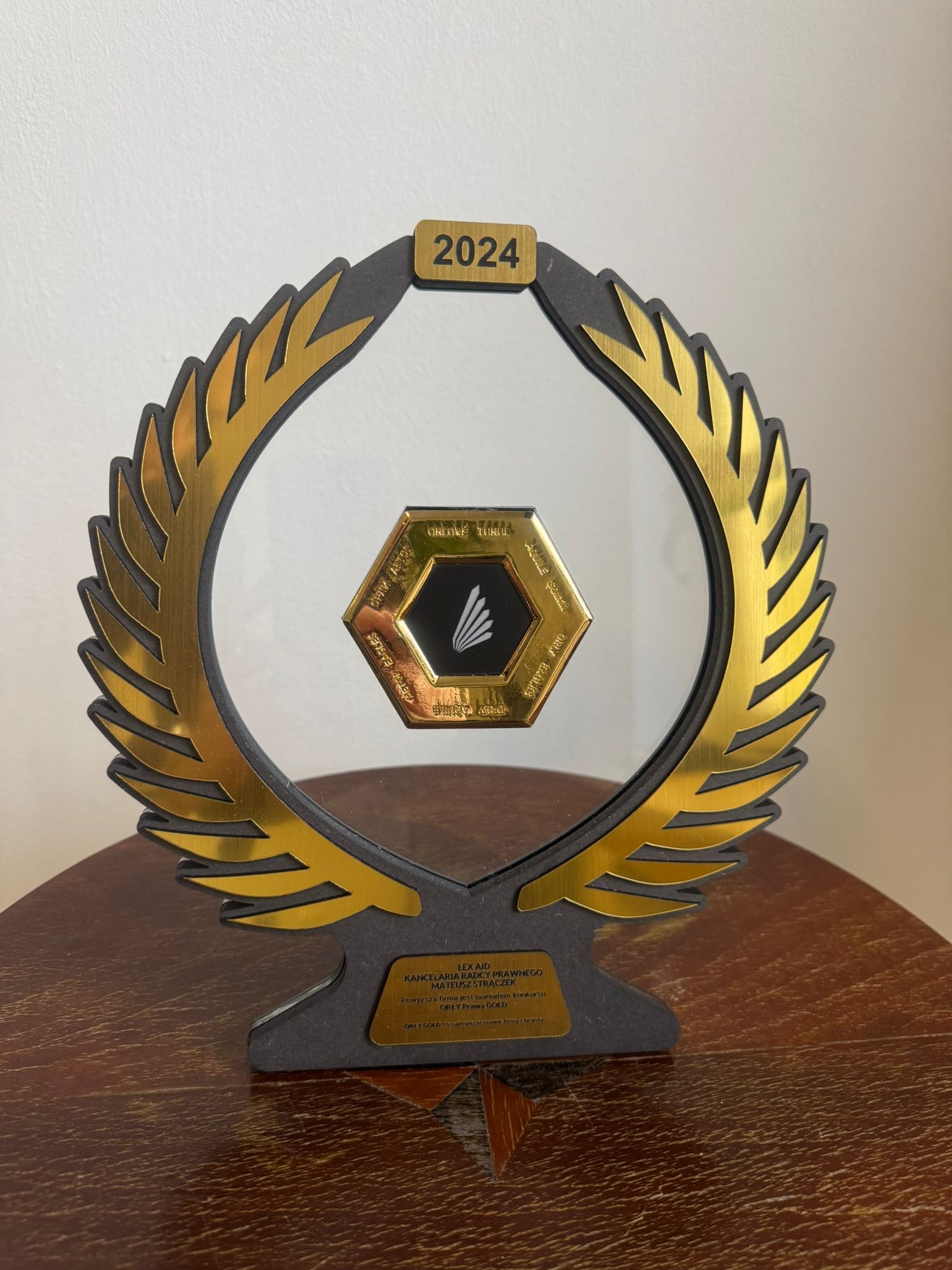 Statuetka z roku 2024, nagroda dla Kancelarii Radcy Prawnego Mateusza Strączka, laureata konkursu Orły Prawa Gold, z elementami w kolorze złotym i czarnym, umieszczona na drewnianym stole.