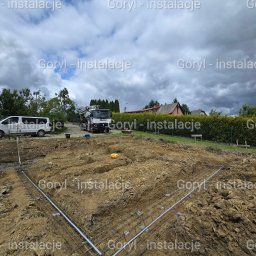 Goryl- instalacje elektryczne - Wykop z ułożonym zbrojeniem i rurami instalacyjnymi, na tle samochód dostawczy i betoniarka. Widok z góry na plac budowy w pochmurny dzień.