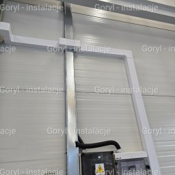 Wiesław Goryl- instalacje elektryczne - Szara ściana z panelami, na której zamontowano białe korytka kablowe, czarną puszkę elektryczną z żółtym znakiem ostrzegawczym i białą skrzynkę z bezpiecznikami.