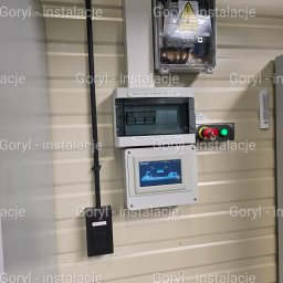 Wiesław Goryl- instalacje elektryczne - Rozdzielnica elektryczna z zabezpieczeniami, sterownikiem i przyciskiem awaryjnym na beżowej ścianie z blachy trapezowej. Widoczna czarna rura instalacyjna.