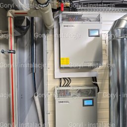 Wiesław Goryl- instalacje elektryczne - Dwa kompensatory mocy biernej Sinexcel zamontowane na ścianie, z widocznymi przewodami i rurami instalacyjnymi. Elementy instalacji elektrycznej i sanitarnej.