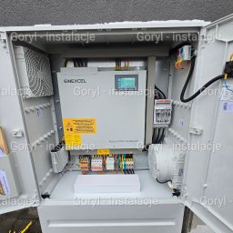 Wiesław Goryl- instalacje elektryczne - Otwarta szafa elektryczna z generatorem SINEXCEL, widoczne okablowanie, bezpieczniki i wentylatory. Szara ściana w tle. Instalacja elektryczna w obudowie.