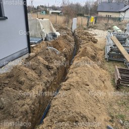 Wiesław Goryl- instalacje elektryczne - Wąski wykop w ziemi z ułożoną niebieską rurą instalacyjną, widok z góry, obok budynku i rusztowania. Etap prac ziemnych przed zasypaniem.