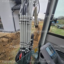 Wiesław Goryl- instalacje elektryczne - Wnętrze kabiny koparki podczas wykopu pod instalacje na działce z widokiem na rów, dom i przyczepę. Perspektywa z kabiny operatora, skupienie na ramieniu koparki.