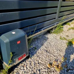 Szara, przesuwna brama wjazdowa z widocznym napędem BFT Ultra, zamontowanym na żwirowym podłożu, z elementami trawy i chwastów.