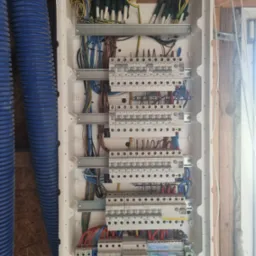 Otwarta skrzynka rozdzielcza z widocznymi bezpiecznikami, przewodami o różnych kolorach i listwami zaciskowymi, zamontowana na ścianie z widoczną strukturą drewna, obok niebieski przewód wentylacyjny.