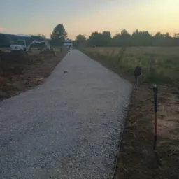 Utwardzona żwirowa droga w perspektywie, z wykoparką w tle, wczesnym rankiem. Widoczne szpadle i wytyczone granice, sugerujące świeżo zakończone prace.